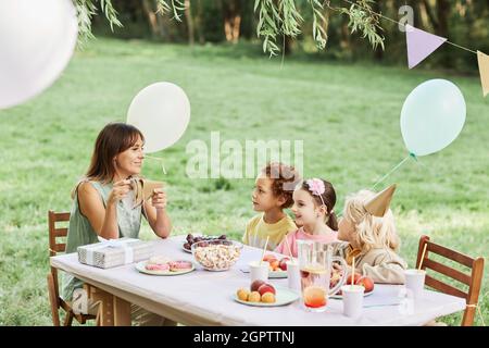 Porträt einer jungen Mutter, die mit einer Gruppe von Kindern während der Geburtstagsfeier im Sommer am Picknicktisch sitzt, Platz zum Kopieren Stockfoto