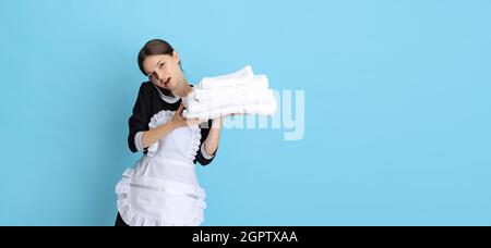 Junge attraktive Zimmermädchen in Uniform mit aufgeklebten frischen Handtüchern, die auf blauem Hintergrund isoliert telefonieren. Flyer Stockfoto