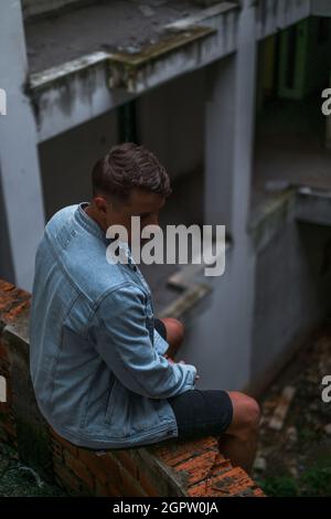 Porträt eines jungen, gutaussehenden kaukasischen Mannes in Denim-Jacke, der auf der verlassenen Baustelle sitzt und nach unten blickt Stockfoto