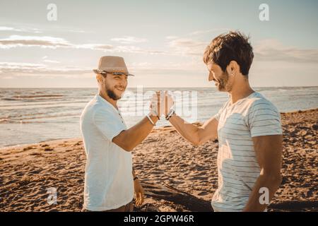 Porträt zweier lächelnder Typen am Strand, die sich die Hände schütteln - Glückliche beste Freunde, die sich gegenseitig mit einem Händedruck begrüßen Stockfoto