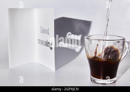 Der Kaffee wird in eine Glastasse gegossen. Die Morgenstrahlen der Sonne scheinen auf einer Tasse Kaffee und einer Karte mit einem guten Morgen Wunsch Stockfoto