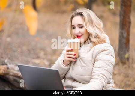 Ein schönes Mädchen in einem Herbstpark hält ein Glas Wegwerfkaffee in der Hand und arbeitet an einem Laptop Stockfoto