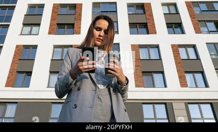 Eine Frau in einem Geschäftsanzug trinkt Kaffee aus einer Plastikbecher und blickt vor dem Hintergrund eines Gebäudes in das Telefon Stockfoto