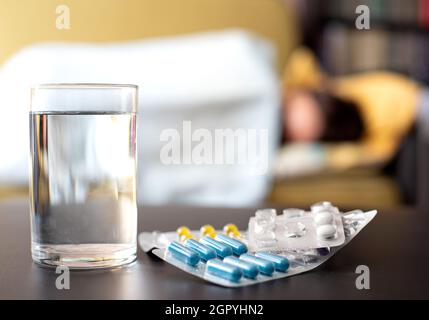 Tabletten und ein Glas Wasser auf dem Tisch im Hintergrund liegt eine kranke Person im Bett Stockfoto