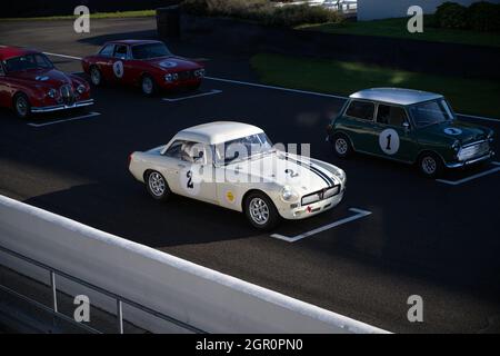 Ein klassischer weißer MGB, der von der Sonne auf dem Startplatz bei Goodwood hervorgehoben wird, und weitere Oldtimer stehen ebenfalls auf dem Startplatz. Stockfoto