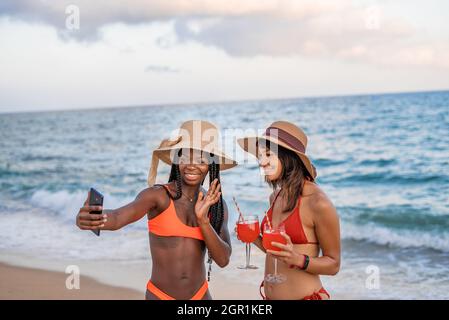 Fröhliche junge afroamerikanische Frau winkt Hand und grüßt, während sie mit ihrer Freundin am Strand in der Nähe des Meeres steht und während des Sommerurlaubs an der tropischen Küste einen Videoanruf über das Smartphone macht Stockfoto