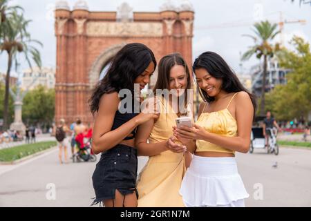 Fröhliche junge, vielfältige Freundinnen in trendigen Outfits diskutieren Fotos auf dem Handy, während sie während der Sommerferien in Spanien in der Nähe des historischen Triumphbogens in Barcelona stehen Stockfoto