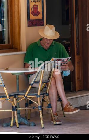 Ein älterer Herr mit panamahut sitzt an einem Tisch außerhalb einer griechischen Taverne und liest an einem Sommertag auf zakynthos zante griechenland eine Zeitung. Stockfoto