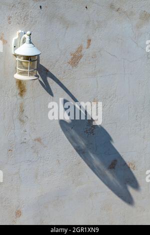 Vintage Außenlampe oder Laterne, die einen langen Schatten auf eine weißgetünchte Wand eines griechischen Gebäudes in der Mittagssonne wirft. Laterne oder Lampe an einer Wand montiert. Stockfoto