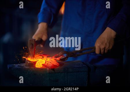 Qualifizierter Schmied in blauen Overalls mit Zange und Hammer, während er mit geschmolzenem Metall arbeitet. Konzept der Handarbeit und Handwerkskunst. Stockfoto