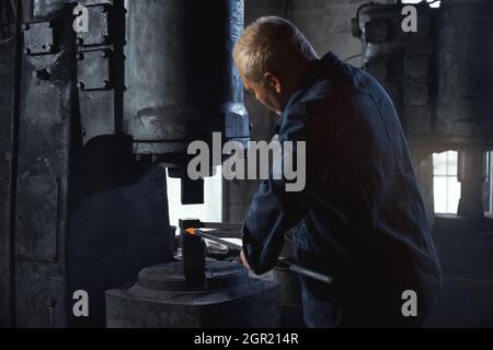 Qualifizierter Schmiedearbeiter trifft in Overalls auf geschmolzenes Metall. Reifer Mann mit schwerem Hammer und Zange für den Arbeitsprozess. Konzept der Schmiedekunst. Stockfoto