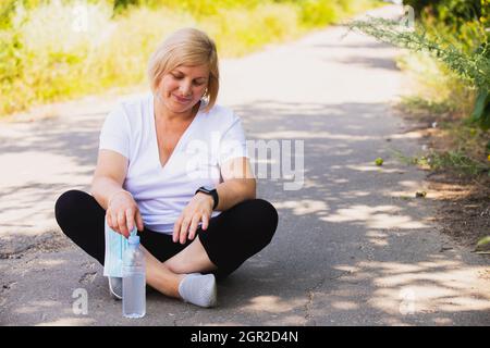 Fitness glückliche Frau sitzt in der Lotusposition auf der Straße im Park Single mit einer Flasche Wasser unten, halten medizinische Maske auf der Hand. Stockfoto