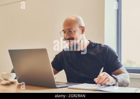 Der junge kaukasische Freiberufler arbeitet an seinem Schreibtisch im Büro mit einem Laptop, während er Notizen macht Stockfoto