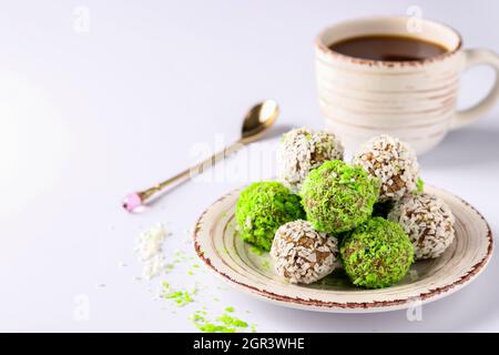 Energiekugeln aus Nüssen, Haferflocken und getrockneten Früchten, bestreut mit grünen und weißen Kokosflocken sowie einer Tasse Kaffee auf weißem Hintergrund Stockfoto