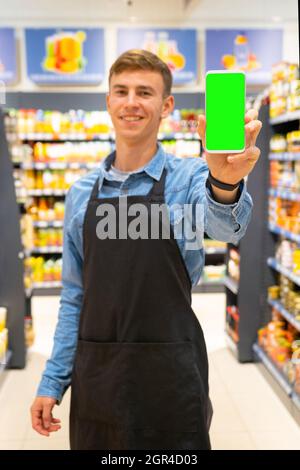 Porträt eines lächelnden Arbeiters in Schürze schwarz im Supermarkt zwischen den Regalen mit den Produkten und zeigt das Smartphone Stockfoto