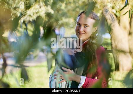 Junge Mutter mit Kind in Babytrage steht im Park. Stockfoto