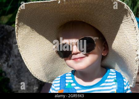 Niedliches kleines Kleinkind, das einen übergroßen Sonnenhut und eine Sonnenbrille trägt, die sich in der Sonne entspannt Stockfoto