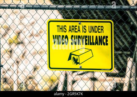 Gelbes Videoüberwachungsschild, das an einem Kettengliederzaun mit schwarzen Schriftzügen befestigt ist und diesen BEREICH LIEST, WIRD VIDEOÜBERWACHT. Stockfoto