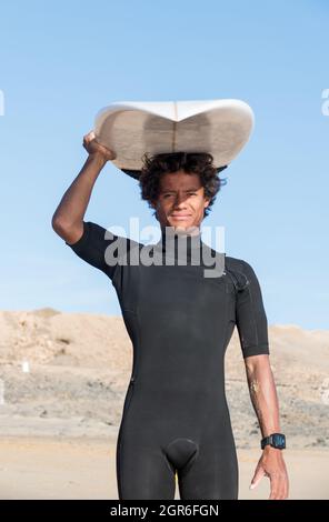 Das junge madagassische afrikanische Surferportrait hält sein Surfbrett und lächelt. Schwarzer kurzhaariger Neoprenanzug. Stockfoto