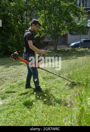 Kovrov, Russland. 28. Juni 2017. Im Innenhof des fünfstöckigen Wohnhauses mäht ein junger Mann das Gras mit einem benzinbetriebenen Unkrauttrimmer Stockfoto