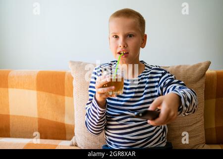 Der junge Mann schaut fern und wechselt die Sender per Fernbedienung. Guy hat sich gelangweilt, was er auf dem fernsehbildschirm sieht. Sitzen Sie auf der Couch in Ihrem Wohnzimmer zu Hause, kopieren Sie Platz. Trinkt Saft aus einem Glas