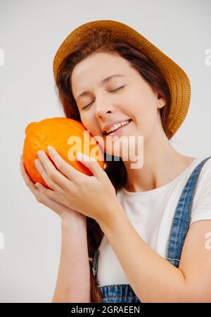 Eine glückliche Frau in Overalls und einem Hut hält einen reifen kleinen Kürbis fest. Stockfoto