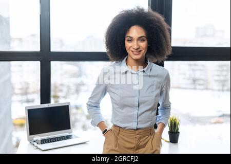 Lächelnde junge Birazialfrau im formellen Anzug blickt auf die Kamera, selbstbewusste Birazialkauffrau steht im Büro und schaut auf die Kamera Stockfoto