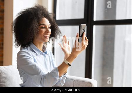 Fröhliche Biracial Frau mit Afro Locken Frisur macht Videoanruf auf dem Smartphone winken, Gruß Freunde oder Kollegen, Mixed-Race-Frau hält Online-Meeting, virtuelle Konferenz am Telefon Stockfoto