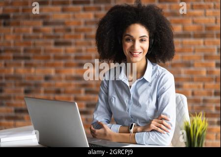 Erfolgreiche Birazialfrau in elegantem Casual-Hemd steht in selbstbewusster Pose mit gekreuzten Armen, selbstbewusste Unternehmerin im Heimbüro blickt auf die Kamera Stockfoto