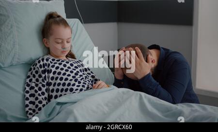 Kranke kleine Tochter mit Sauerstoff-Nasenschlauch im Bett liegend schlafen während der Genesung Untersuchung in der Krankenstation. Besorgte Mutter, die neben ihr betet und auf eine medizinische Behandlung wartet Stockfoto