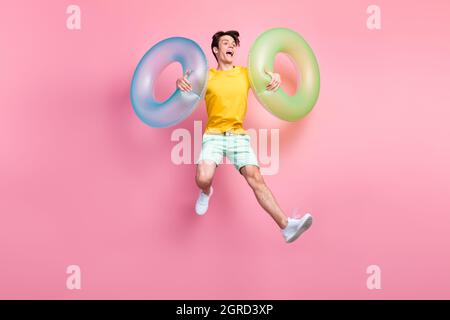 Foto von lustigen positiven Kerl springen tragen aufblasbare Kreise tragen gelbe T-Shirt Shorts Sneakers isoliert auf rosa Hintergrund Stockfoto