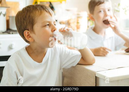 Ein Junge trinkt Milch aus einer Flasche in der Küche zu Hause. Frühstück mit Milch. Glückliches Kind in einem weißen T-Shirt trinkt Milch aus einer Flasche Stockfoto