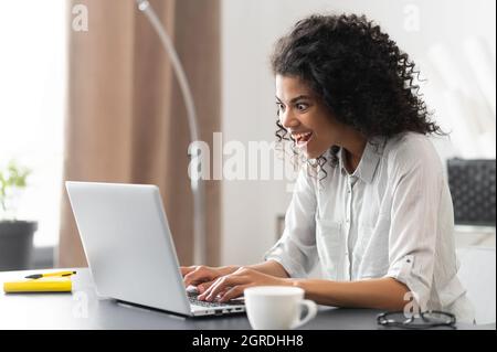 Eine afroamerikanische Geschäftsfrau in einem legeren Hemd, die am Schreibtisch saß und auf den Laptop-Bildschirm starrte, überraschte eine weibliche Büroangestellte mit gemischter Rasse, die unerwartete gute Nachrichten las Stockfoto
