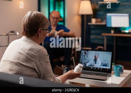 Älterer Patient mit einer Flasche Pillen in der Hand, der eine Videokonferenz auf dem Laptop für einen Termin mit dem Arzt zu Hause verwendete. Pensionierte Frau bekommt Medikamentenpräsition auf Online-Fernuntersuchung Stockfoto