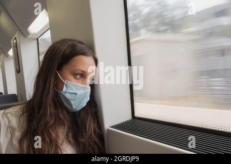 Hübsches Mädchen mit langen Haaren, das in einem Zug vor dem Fenster sitzt. Ein junger Zugpassagier mit Schutzmaske. Sie scheint zu reisen Stockfoto