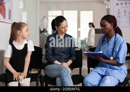 Familie diskutiert medizinische Behandlung mit afroamerikanischen Krankenschwester im Krankenhaus Warteraum während klinischer Termin. afroamerikanische Krankenschwester schreibt klinische Expertise. Support-Service im Gesundheitswesen Stockfoto