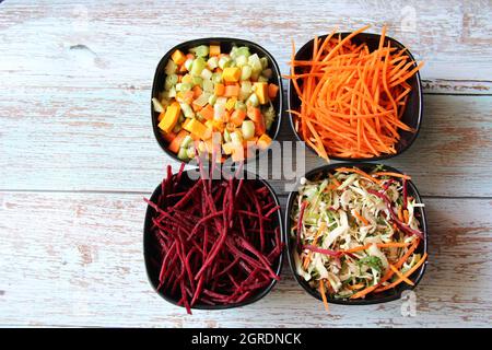 Gemüse, Bio, roh, sehr klein geschnitten und gerieben, wie Karotten, Rüben, unter anderem, in vier schwarzen Schalen auf einem alten Holztisch. Obere Ansicht. Stockfoto