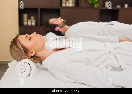 Seitenansicht einer Frau im Bademantel, die auf dem Massageliegen neben einem verschwommenen Freund im Spa-Center lag Stockfoto
