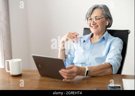 Aufmerksame ältere weibliche Büroangestellte Geschäftsfrau in Brille sitzt am Schreibtisch, im Internet surfen, hören einen Podcast, E-Mails von den Kunden, mit Entdeckung neue Unternehmens-App Stockfoto