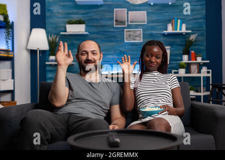Interrassisches Paar winkt bei Videoanruf-Kamera mit Technologie für Online-Fernkommunikation im Internet. Multiethnische Partner, die zu Hause auf einer Videokonferenz mit Freunden sprechen Stockfoto