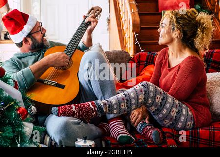 Paar zu weihnachten zu Hause feiern und Spaß haben zusammen mit Lächeln und Spaß - Mann spielen eine Gitarre und Frau schauen ihn an. Liebe und Freizeit in der Halle Stockfoto