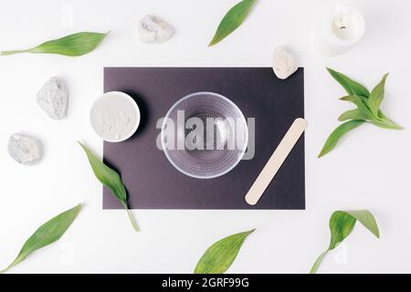 Draufsicht auf leere Glasschüssel für die Herstellung von natürlichen hausgemachten kosmetischen Maske aus Ton zwischen grünen Blättern auf weißem Tisch Stockfoto