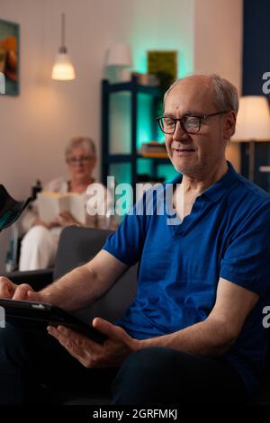 Nahaufnahme eines älteren Mannes, der ein digitales Tablet auf dem Sofa im Wohnzimmer hält und anschaut. Rentner mit modernem Gadget zu Hause und ältere Frau im Hintergrund sitzt im Rollstuhl. Stockfoto