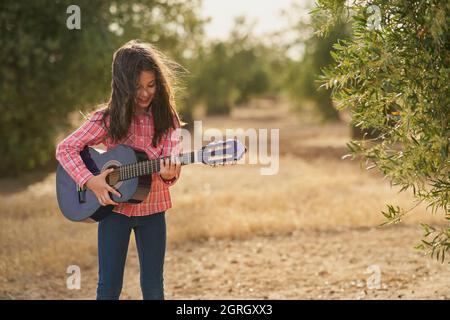 Brunette Mädchen spielt ihre Gitarre glücklich bewegen ihre Haare in einem Feld von Olivenbäumen bei Sonnenuntergang Stockfoto