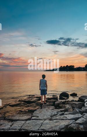 Rückansicht eines Kindes am Seeufer, das den Himmel bei Sonnenuntergang betrachtet. Stockfoto