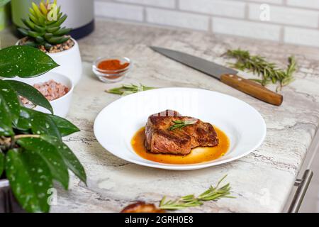 Frisch zubereitetes, geröstetes Rindersteak mit Gewürzen und Rosmarin auf weißem runden Teller auf dem Küchentisch im Hintergrund. Stockfoto