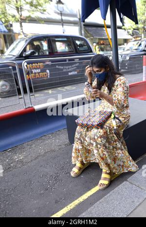 London, England, Großbritannien. Junge Frau in einem blumigen Kleid mit COVID-Maske und Mobiltelefon Stockfoto