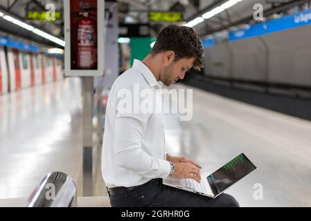 Seitenansicht eines Mannes in eleganter Freizeitkleidung, der auf der Bank sitzt und auf der Netbook-Tastatur tippt, während er auf der U-Bahn-Station auf den Zug wartet Stockfoto