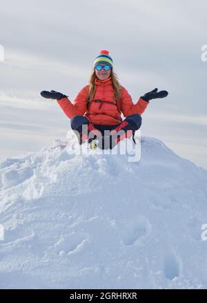 Die Wandererin in warmer Winterkleidung sitzt auf dem Schneehaufen, ihre Arme sind ausgestreckt und ihre Beine gekreuzt, glücklich und lächelnd. Gipfel, Spitze, goa Stockfoto