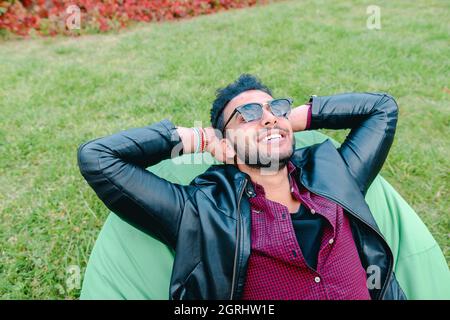 Porträt eines jungen arabischen Mannes, Student liegt auf einem Stuhl, lächelt, schaut in den Himmel, meditiert über den Hintergrund des grünen Grases im Garten im Freien. Stockfoto
